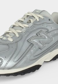 Ασημί αθλητικό παπούτσι New Balance με λευκά κορδόνια, υφασμένους πλαϊνούς πίνακες και μαύρες λεπτομέρειες στη σόλα.