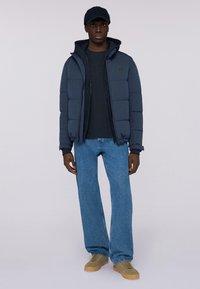 Námorně modrá péřová bunda na zip, nošená přes tmavý top, v kombinaci se světle modrými džíny rovného střihu a béžovými teniskami.