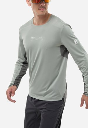 Homme portant une chemise Millet à manches longues gris clair avec des panneaux gris foncé, un pantalon noir et des lunettes de soleil teintées orange, posé devant un fond blanc.