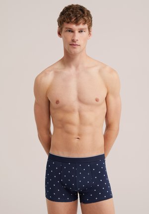 Jeune homme aux cheveux bouclés portant un boxer bleu marine à motifs, debout devant un fond clair uni.