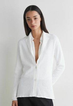 Kvinde med langt mørkt hår iført en hvid cardigan med knapper over en hvid skjorte, stående mod en ensfarvet lys væg.