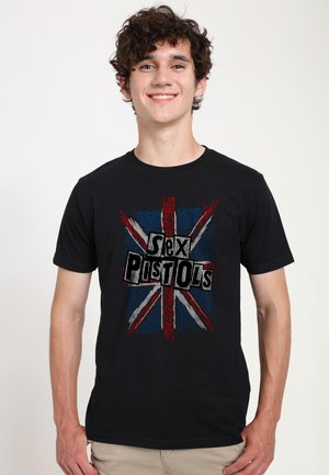 Giovane uomo sorridente che indossa una maglietta nera con una bandiera Union Jack usurata e il testo "Sex Pistols" sul davanti.