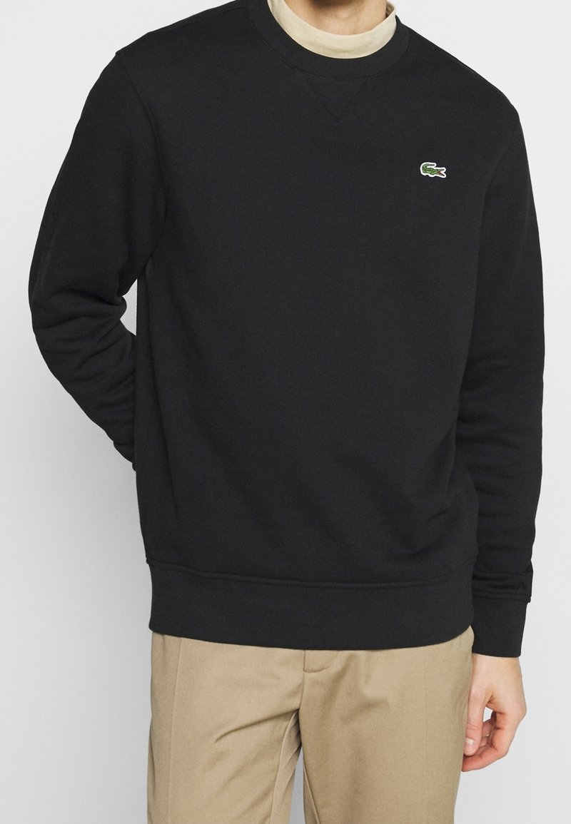 Homme portant un sweat-shirt noir avec un petit logo crocodile vert sur la poitrine, pantalon beige et chemise beige en dessous, debout devant un fond blanc.