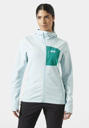 Kobieta ubrana w jasnoniebieską kurtkę z suwakiem, z morskim zielonym kieszonką na piersi i czarnymi spodniami, stojąca na jednolitym białym tle.