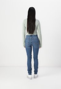 Svijetlozelena majica dugih rukava u kombinaciji s uskim plavim trapericama i bijelim tenisicama, prikazana s leđa na jednostavnoj bijeloj pozadini.