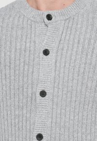 Grå ribbestrikket cardigan med rund krage, fem svarte knapper og et mykt, teksturert strikket stoff. Nærbilde av fronten.