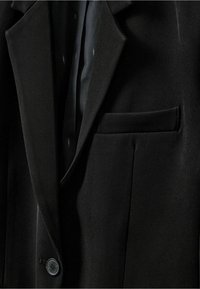 Sacou negru cu o textură netedă, cu rever în fanteză, închidere cu un singur buton și un buzunar pe pieptul stâng. Interiorul căptușit cu un material închis la culoare.