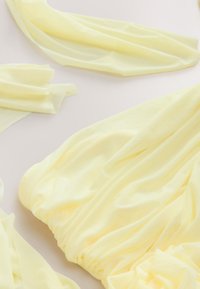 Pehmeitä, virtaavia vaaleankeltaisia kangaspaloja litteästi aseteltuna, joissa on hellävaraisia taitoksia ja kevyitä varjoja neutraalilla taustalla.