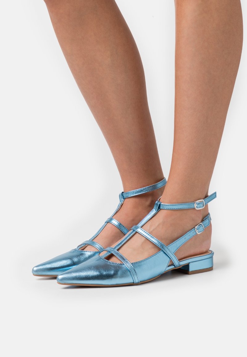 Ballerines métalliques bleu avec des pointes pointues, des sangles croisées et des boucles à la cheville. Talons plats pour un meilleur confort. Texture lisse avec une finition brillante.