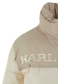 Béžová páperová bunda s vysokým golierom a jemnou textúrou. Na hornej časti hrude je vyšívané slovo "KARL" v bielej farbe.