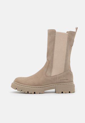 Bottines beiges en suède avec un panneau en maille texturée, bout arrondi, semelle épaisse en caoutchouc et détail de marque sur le côté.