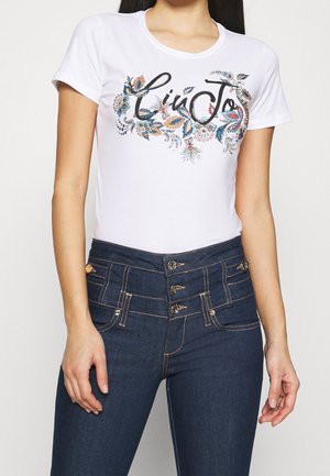 Frau trägt weißes T-Shirt mit kurzen Ärmeln mit Blumenstickerei und dem Schriftzug "Liu Jo", kombiniert mit hoch taillierten dunkelblauen Jeans mit mehreren Knöpfen.