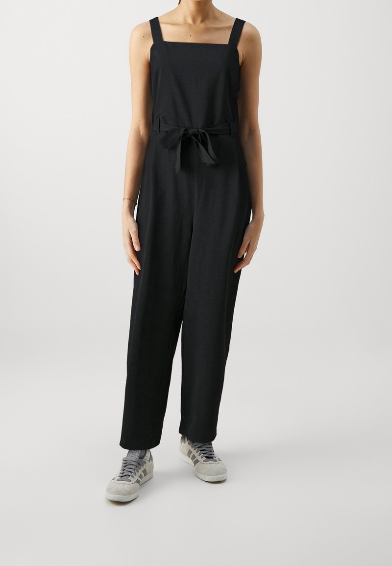Femme portant une combinaison noire sans manches avec une ceinture nouée à la taille et des baskets blanches, debout devant un fond clair uni.