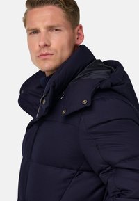 Tmavě modrá puffer bunda s vysokým límcem, lesklou černou kapucí a zlatými patentkami. Strukturální látka se šitým prošívaným vzorem.