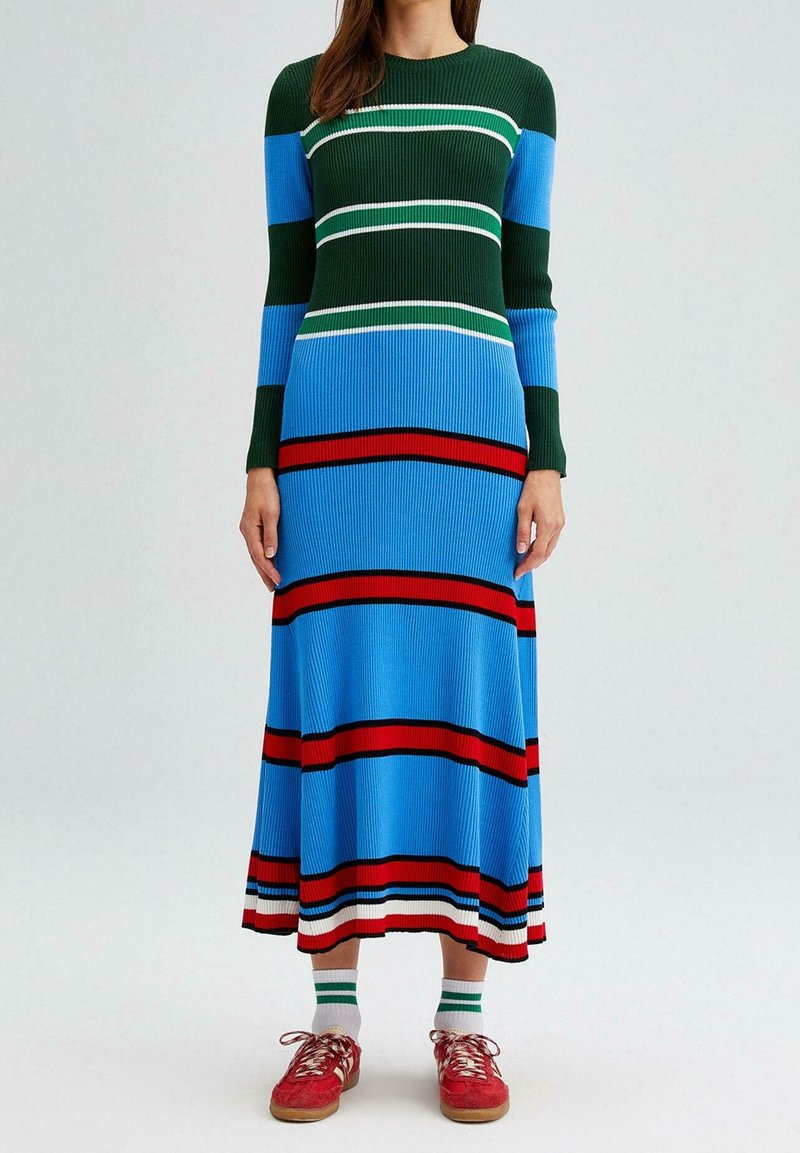 Femme portant une longue robe en maille côtelée avec des rayures horizontales vertes, bleues, rouges, blanches et noires, ainsi que des baskets rouges avec des chaussettes blanches.