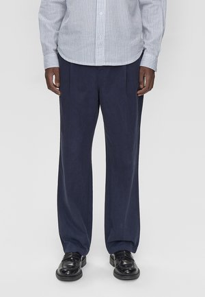Man wearing navy wide-leg trousers, black leather shoes, and a striped button-up shirt standing against a plain background.