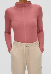 Pitkähihainen bodysuit pehmeässä vaaleanpunaisessa kankaassa, jossa on korkea kaulus ja etusauma. Yhdistetty beigeihin nyöripökiäisiin, joissa on sivutaskut.