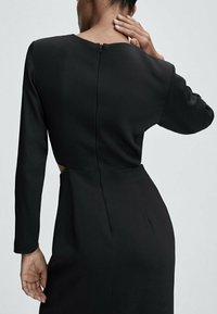 Svart klänning med långa ärmar, rund hals och dragkedja i ryggen. Har en utskärning vid midjan som framhäver eleganta linjer och en mjuk textur.