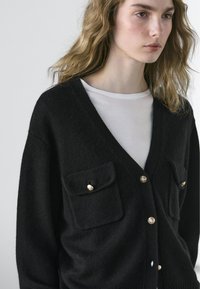 Zwarte gebreide cardigan met een diepe V-hals, twee zakken aan de voorkant met klep en goudkleurige knopen. Zachte textuur en een ontspannen pasvorm.