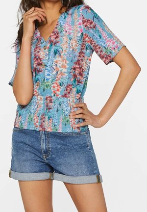 Woman wearing a blue floral V-neck blouse with buttons and elastic waist, paired with rolled-up denim shorts.