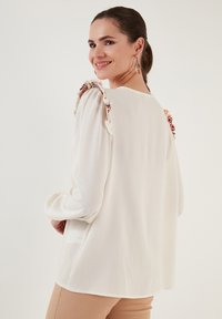 White long-sleeve blouse with ruffled shoulders and colorful embroidered accents. Soft fabric with a relaxed fit, round neckline, and subtle sheen.