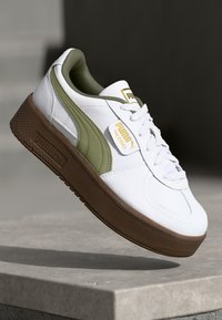 Zapatilla de piel blanca con acentos verdes, suela de goma texturizada marrón, diseño de cordones y detalles en dorado en la lengüeta.