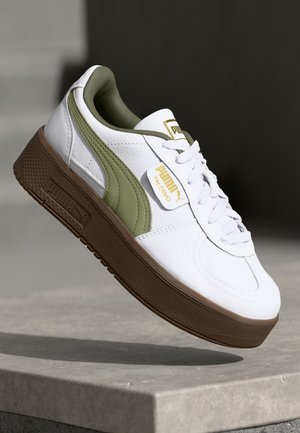 Zapatilla de piel blanca con acentos verdes, suela de goma texturizada marrón, diseño de cordones y detalles en dorado en la lengüeta.