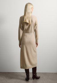 Vestido de punto beige con mangas largas, cintura ajustada y largo midi. Combinado con botas altas de color burdeos. Textura suave.