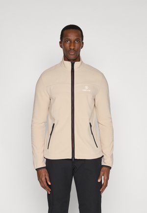 Mand iført en beige zip-up jakke med sorte lynlåsdetailler og sorte bukser, stående mod en ensfarvet hvid baggrund.
