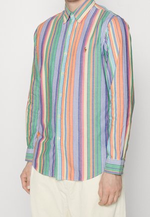Homme portant une chemise à manches longues boutonnée avec des rayures verticales en vert, orange, bleu, violet et blanc, associée à un pantalon de couleur claire.