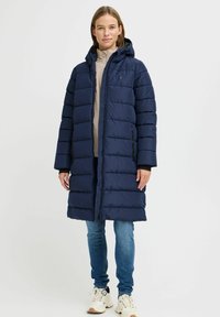 Marineblauer Puffermantel mit Kapuze, Reißverschluss, gestepptem Muster und zwei seitlichen Taschen. Kombiniert mit hellblauen Jeans und Sneakers.
