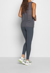 Šedý top bez rukávů spárovaný s tmavě modrými legínami. Obuv bílé barvy se stříbrnými a neonovými akcenty. Důraz na střih a atletický styl.