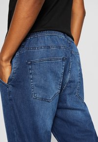 Džínové kalhoty s elastickým pasem v tmavě modré barvě, s kapsami na zádech zdobenými prošitím a hladkou texturou.