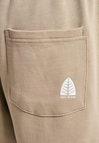 Beige szövetzseb, varrott varratokkal, fehér levél logóval és alatta a "JUST RHYSE" szöveggel. Puha tapintású, laza dizájn.