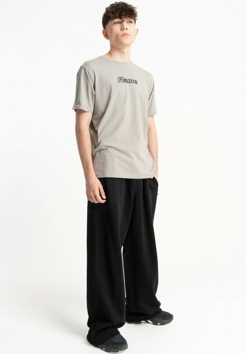 Młody mężczyzna stoi w szarej koszulce "Flame", luźnych czarnych spodniach i czarnych trampkach na białym tle.