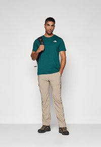 Teal kortermet T-skjorte med en liten logo, beige cargobukser med sidelommer, og svarte tursko med oransje detaljer, supplert med en ryggsekk.