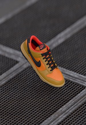 Ein einzelner orange-gelber Nike-Sneaker mit schwarzem Swoosh und schwarzen Schnürsenkeln, der auf einer dunklen Metallgitteroberfläche ruht.