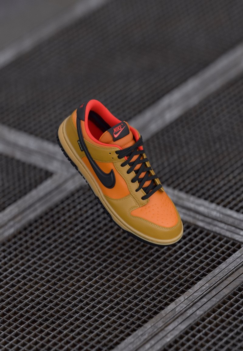 Une basket Nike orange et jaune avec un swoosh noir et des lacets noirs, reposant sur une surface en grille métallique sombre.