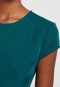Teal lyhythihainen paita pyöreällä kaula-aukoilla, valmistettu pehmeästä kankaasta. Ominaisuuksina minimaalinen ompelu ja sileä pinta.
