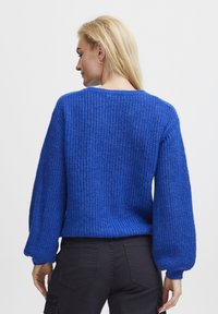 Maglione azzurro lavorato a maglia con una trama testurizzata, maniche a palloncino e scollatura rotonda, abbinato a pantaloni scuri, visto da dietro.