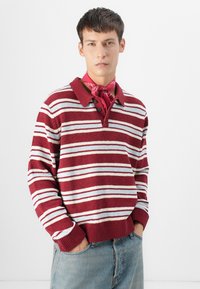 Pull polo tricoté rouge et blanc à rayures avec un col, comportant trois boutons au niveau du cou ; porté avec un foulard rose et un jean bleu clair.