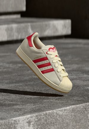 Witte en rode sneaker met drie rode strepen en veters, zwevend boven een grijs betonnen oppervlak met trappen op de achtergrond.