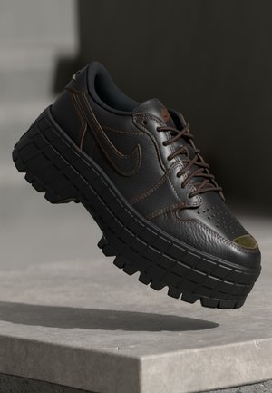 Mustavärinen Nike-nahkainen lenkkari, jossa paksu, kuvioitu pohja ja ruskea ompelu, leijuu harmaan betonipinnan yllä.