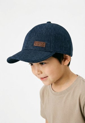 Young boy wearing a dark blue fabric baseball cap with a brown "Stern Taler" label, smiling and looking slightly downward.