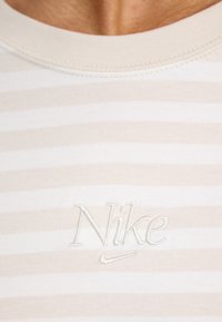 Juostuota marškinėliai smėlio ir šviesiai pilkos spalvos, su tekstūrine išsiuvinėta Nike logotipu kairėje krūtinės pusėje. Minkšta medvilnė, apvalaus kaklo dizainas.