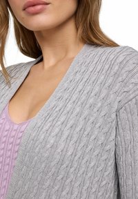 Grauer, gestrickter Cardigan mit V-Ausschnitt, der aus strukturiertem Stoff besteht, langen Ärmeln und über einem helllila Oberteil getragen wird.