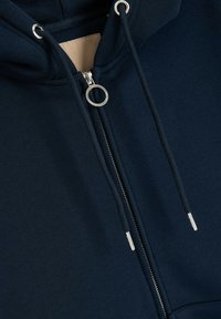 Granatowa bluza z zamkiem błyskawicznym, wykonana z miękkiego materiału; wyposażona w srebrne elementy, troczki i pętelkę do suwaka. Gładka tekstura z dopasowanym krojem.