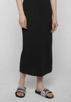 Personne portant une longue robe noire et des sandales à enfiler noires avec des paillettes argentées et un texte noir en gras sur les brides.