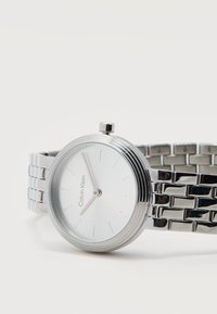 Ασημένιο ρολόι χειρός Calvin Klein με μινιμαλιστικό λευκό καντράν, χωρίς αριθμούς, και συνδεδεμένο μεταλλικό μπρασελέ, δείχνοντας ώρα 12:58.