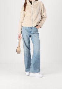 Faux fur cream jacket with a high collar, paired with light blue wide-leg jeans and white sneakers, holding a beige handbag.